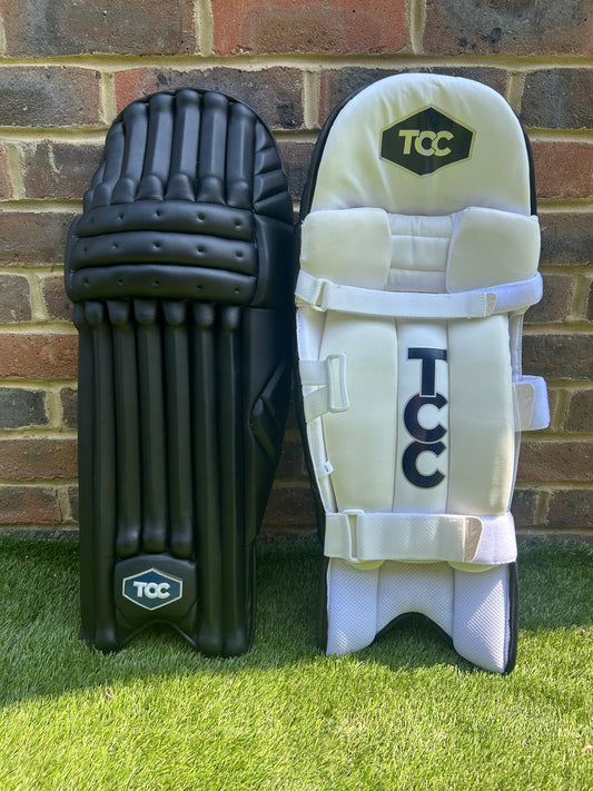 TCC Premium Black Batting Pads with Navy Logo for front and inside view positioned on grass against a brick wall