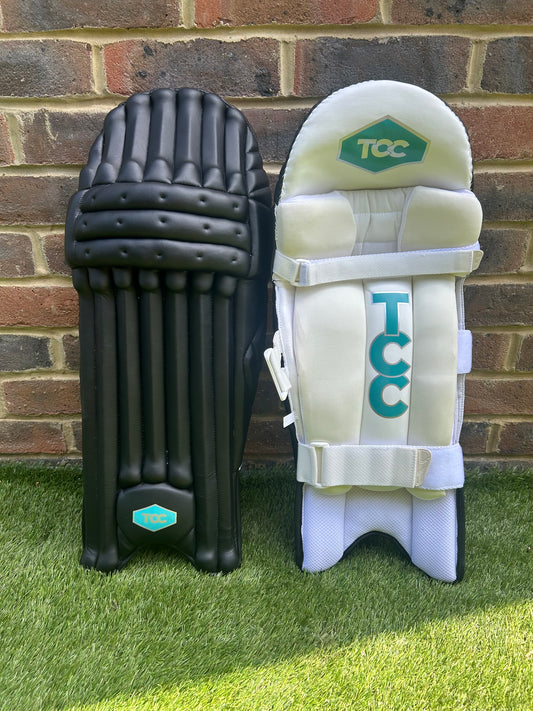 TCC Premium Black Batting Pads with Teal Logo positioned on the grass leaning against a brick wall