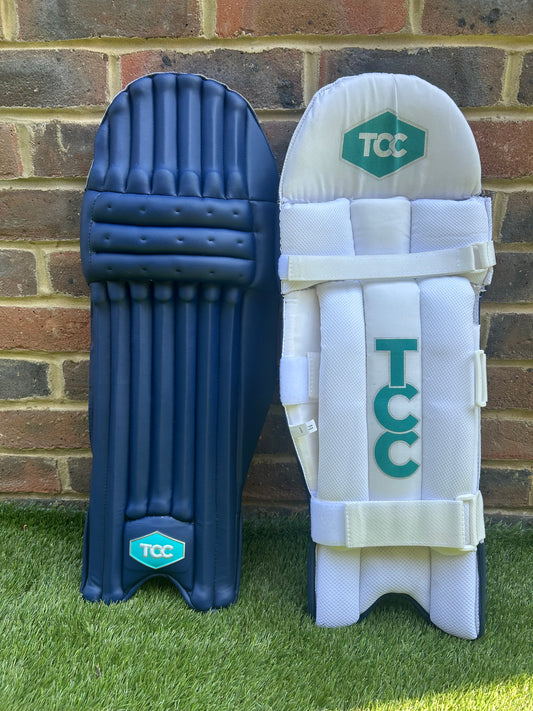 TCC Youth Navy Batting Pads with Teal Logo positioned on the grass against a brick wall showing the front and inside leg of the pads