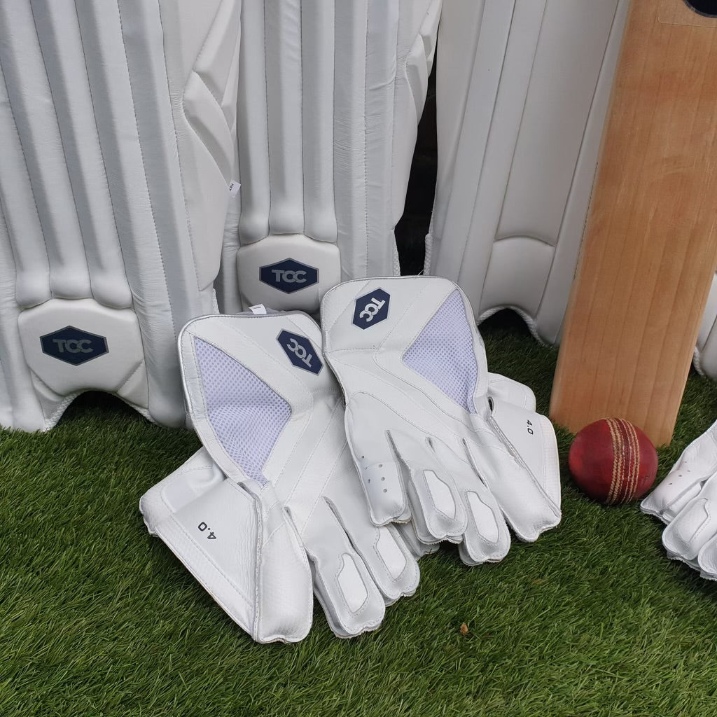 TCC Wicket keeping gloves laid on grass next to pads bat and ball