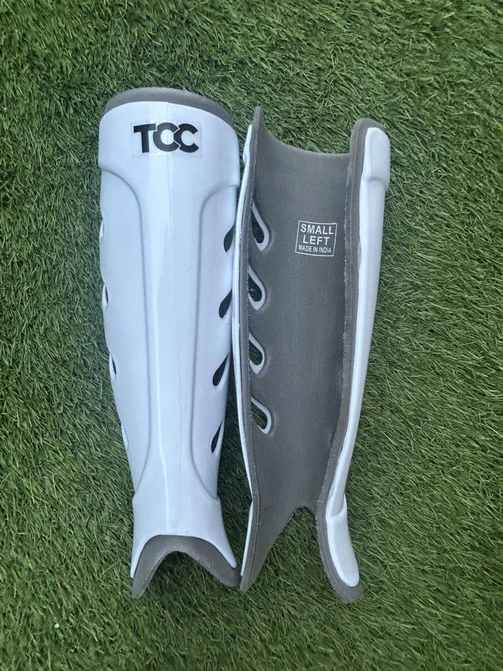 White shin pads with 'TCC' branding on a grassy background, showing the front and reverse view of the shin pads.