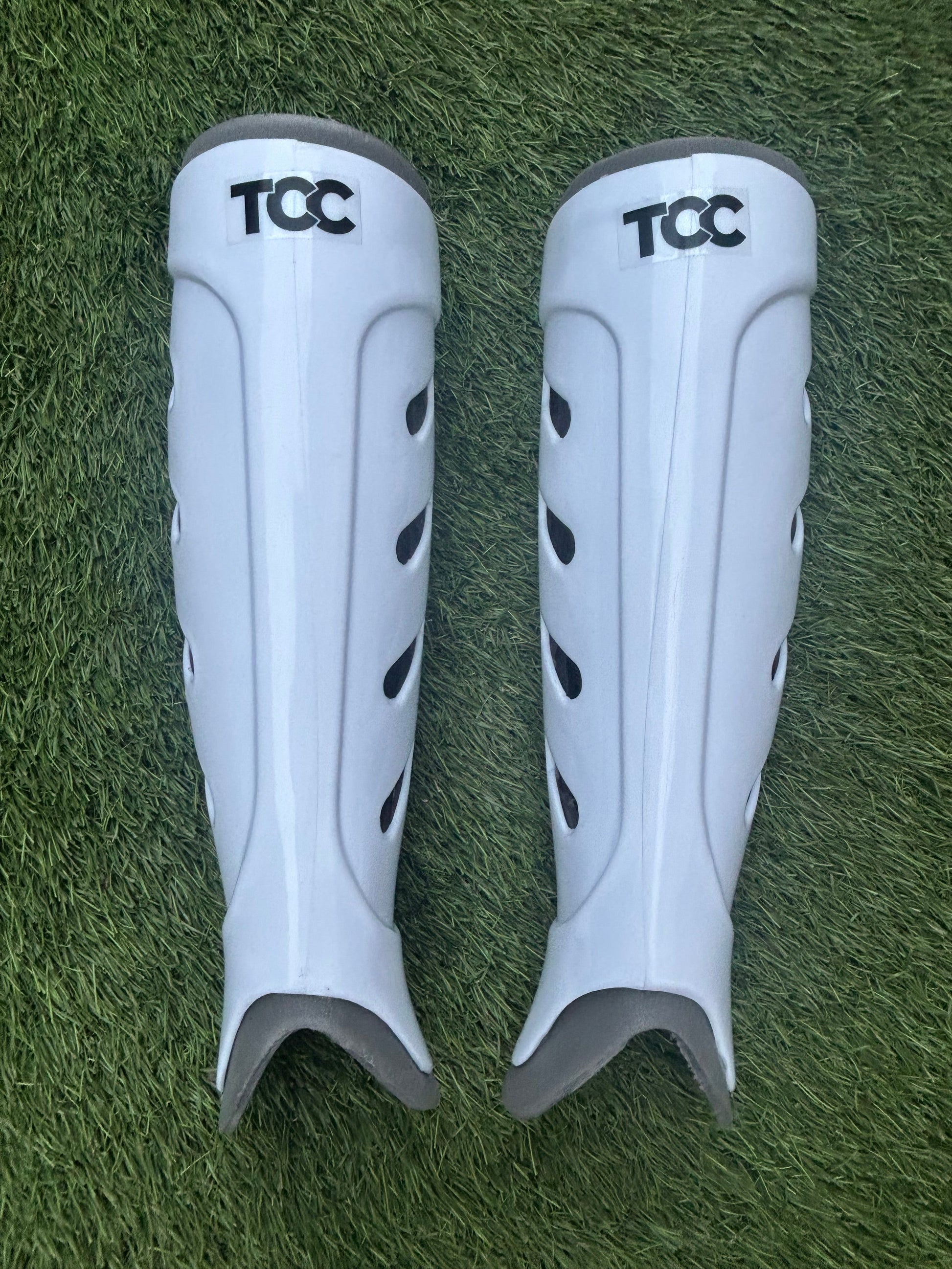 Pair of white shin guards with 'TCC' branding on a grass background