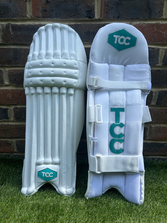 TCC Youth White Batting Pads with Teal Logo positioned on the grass against a brick wall showing the front on and inside leg view of the pads