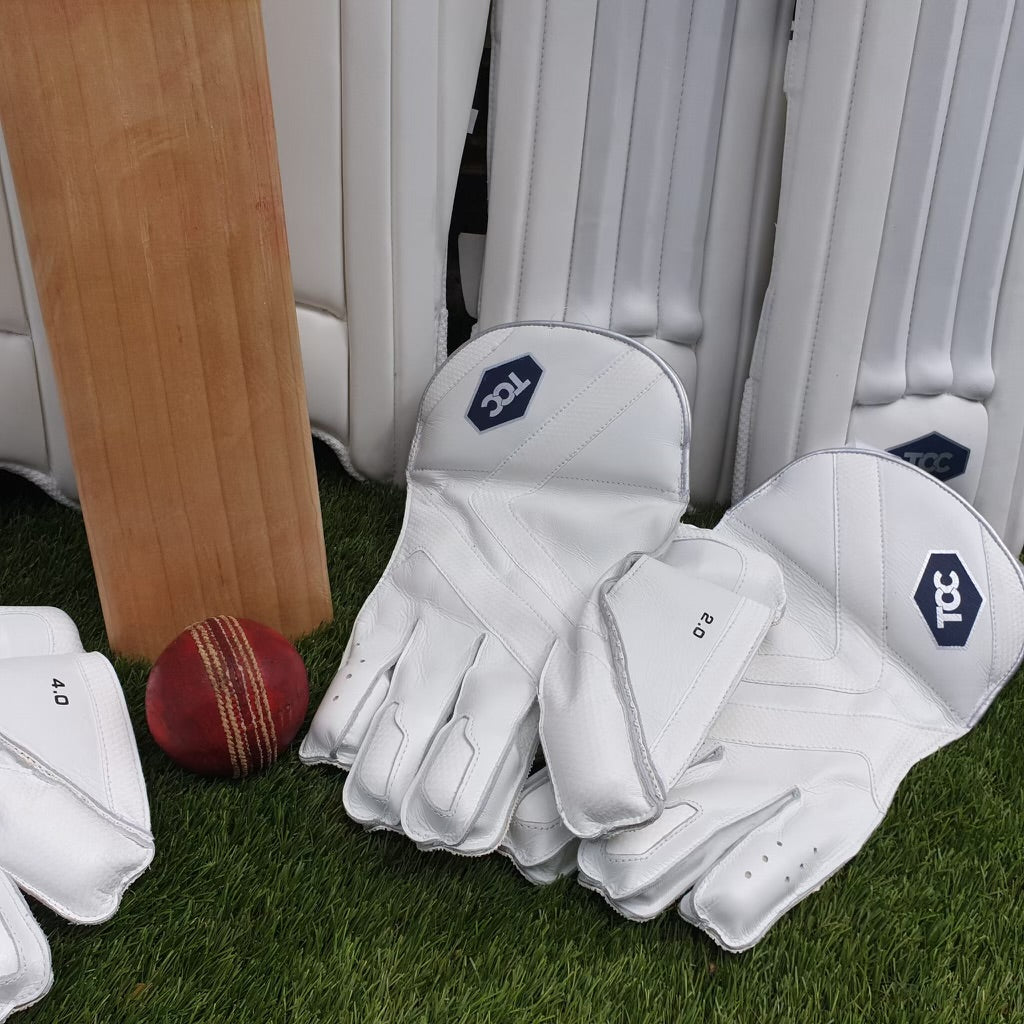 TCC Wicket keeping gloves showing the back of the hand designs alongside pads, bat and cricket ball